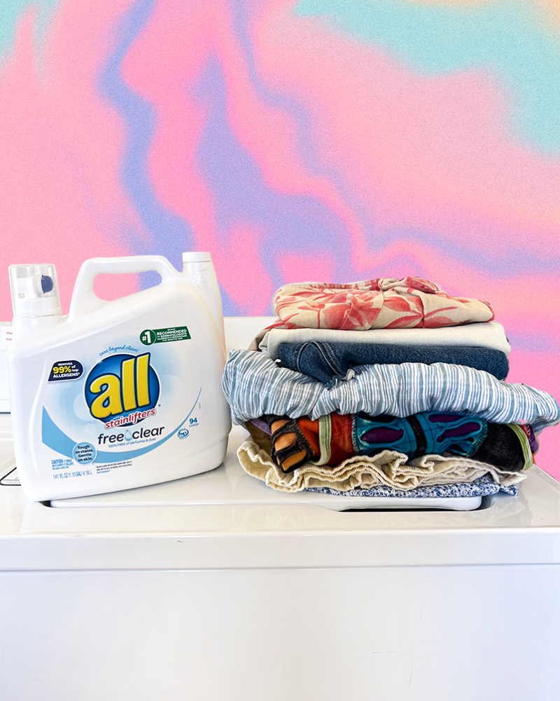A pile of clothes and large bottle of All brand laundry detergent sit on top of a washing machine