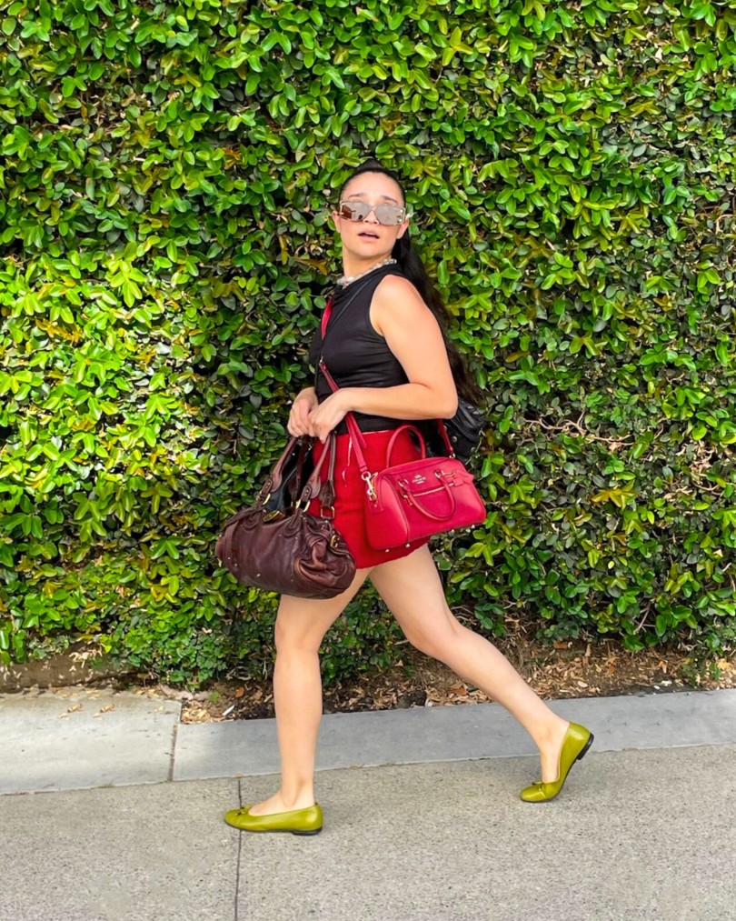 Person walking down a sidewalk wearing a black tank top, red mini skirt and olive ballet flats while carrying several designer purses from Chloe and Coach